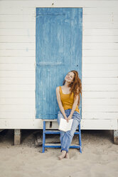 Redheaded woman sitting in front of beach cabin, reading a book - KNSF04330