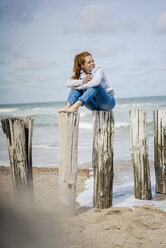 Frau sitzt auf einem Zaun am Meer und trinkt Tee - KNSF04339
