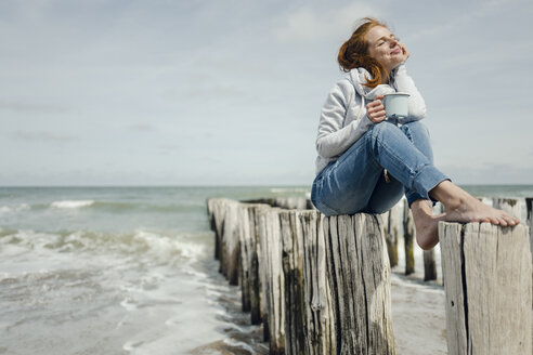 Frau sitzt auf einem Zaun am Meer und trinkt Tee - KNSF04346