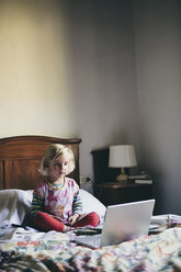Ein dreijähriges Mädchen, das auf einem Bett in einem Hotelzimmer sitzt und aufmerksam auf den Bildschirm eines Laptops schaut. - MINF01814