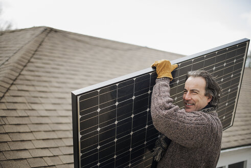 Ein Mann trägt ein großes Solarmodul über einen Bauernhof. - MINF01847