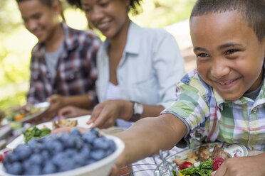 Ein Familienpicknick in einem schattigen Waldstück, Erwachsene und Kinder sitzen am Tisch. - MINF01861