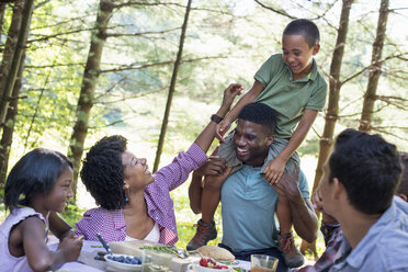 Ein Familienpicknick im Schatten hoher Bäume, ein kleiner Junge sitzt auf den Schultern seines Vaters. - MINF01867