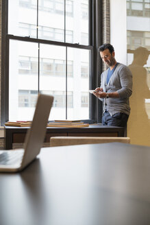 Büroalltag: Ein Mann steht in einem Büro am Fenster und überprüft sein Smartphone. - MINF01892