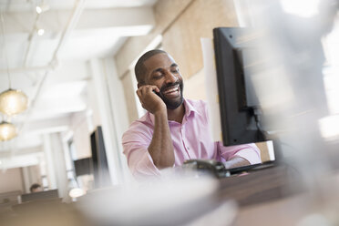 Office life. A man seated at a desk with a computer monitor, using a smart phone. - MINF01898