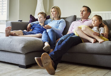 A family of four people, parents and a girl and boy, seated on the sofa together, watching television. - MINF01974