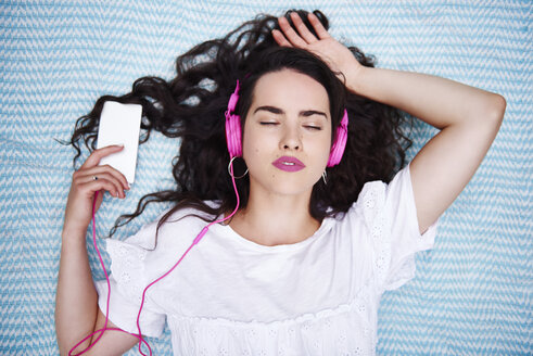 Porträt einer jungen Frau, die auf einer Decke liegt und mit einem Smartphone und rosa Kopfhörern Musik hört - ABIF00750