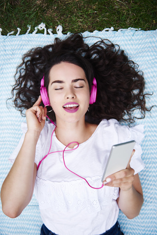 Porträt einer jungen Frau, die auf einer Decke liegt und mit einem Smartphone und rosa Kopfhörern Musik hört, lizenzfreies Stockfoto