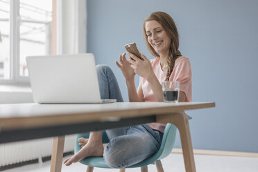 Woman at home sitting at desk with laptop, using smartphone - JOSF02309