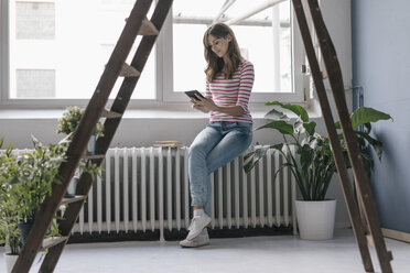 Frau sitzt auf dem Heizkörper in ihrer neuen Wohnung und liest ein E-Book, umgeben von Pflanzen - JOSF02369