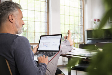 Mature man working from his home office with feet up, using laptop - FKF03041