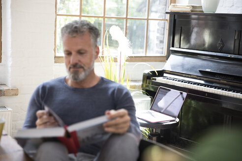 Laptop auf einem Hocker vor einem Klavier, Mann liest in einem Buch - FKF03050