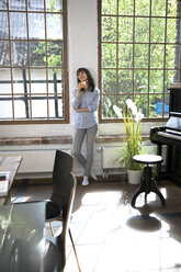 Frau steht am Fenster ihrer Loftwohnung und trinkt Kaffee - FKF03053