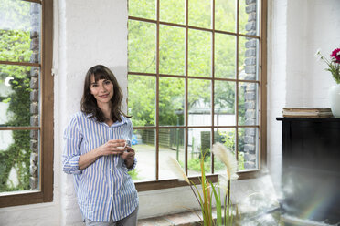 Woman standing at window of her loft apartment, drinking coffee - FKF03057