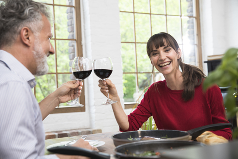 Glückliches Paar sitzt in der Küche, stößt mit Rotwein an und genießt das Abendessen, lizenzfreies Stockfoto