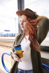 Portrait of redheaded woman with coffee to go and smartphone waiting at platform - ABIF00761