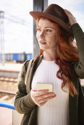 Porträt einer rothaarigen Frau mit Hut und Smartphone am Bahnsteig - ABIF00763
