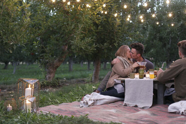 Apple orchard. Couple sitting on the ground, kissing, food and drink on a table. - MINF02300