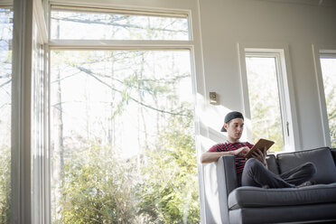 Man wearing a baseball cap backwards, sitting on a sofa, looking at a digital tablet. - MINF02317