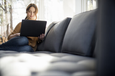 Frau sitzt auf einem Sofa und schaut lächelnd auf ihren Laptop. - MINF02320