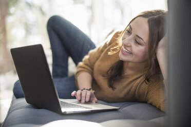 Woman lying on a sofa looking at her laptop, smiling. - MINF02323