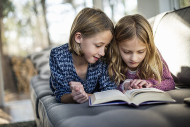 Zwei blonde Mädchen liegen auf einem Sofa und schauen in ein Buch. - MINF02324