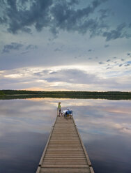 A couple, man and woman sitting at the end of a long wooden dock reaching out into a calm lake, at sunset. - MINF02379