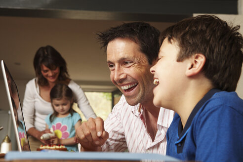 Eine Familie, zwei Eltern und zwei Kinder, die zusammen zu Hause sind, ein Vater und ein Sohn an einem Laptop, eine Mutter und eine Tochter, die einen Kuchen glasieren. - MINF02497