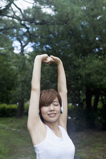 Eine Frau mit Kopfhörern in einem Park in Kyoto, die eine Joggingausrüstung trägt und sich vor dem Training dehnt. - MINF02526