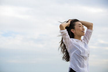 Woman stands in front of a cloudy sky and reaches into her hair - MINF02569