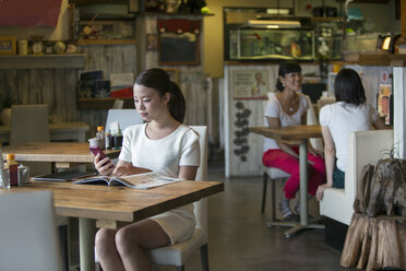 Woman sitting at a table in a cafe, looking at her mobile phone, two women at a table in the background. - MINF02619