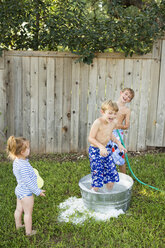 Drei Kinder spielen in einem Garten mit einer mit Wasser gefüllten Wanne und einem Schlauch. - MINF02639