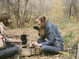 Ein lächelndes Paar sitzt im Wald auf dem Boden und trinkt Kaffee. - MINF02698