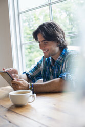 Ein Mann sitzt mit einer Tasse Kaffee in einem Café und benutzt ein digitales Tablet. - MINF02769