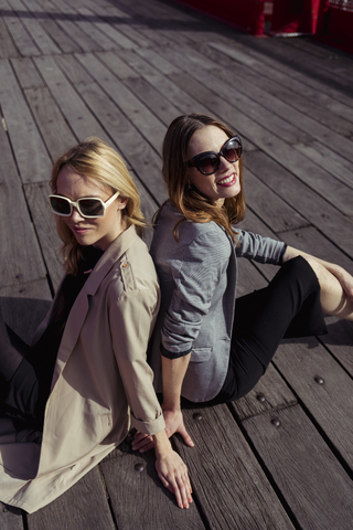 Porträt von zwei modischen jungen Frauen mit Sonnenbrillen, die auf einem Holzboden sitzen, lizenzfreies Stockfoto