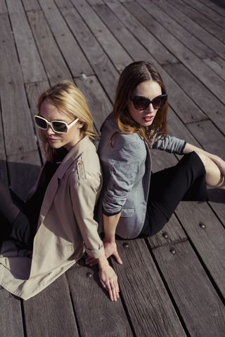 Porträt von zwei modischen jungen Frauen mit Sonnenbrillen, die auf einem Holzboden sitzen, lizenzfreies Stockfoto