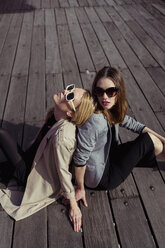 Portrait of two fashionable young women sitting on wooden floor wearing sunglasses - MAUF01530