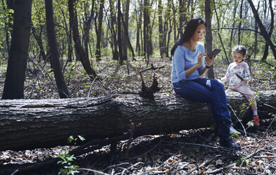 Mother and daughter in park,sitting on tree trunk, using smartphone - AZF00020