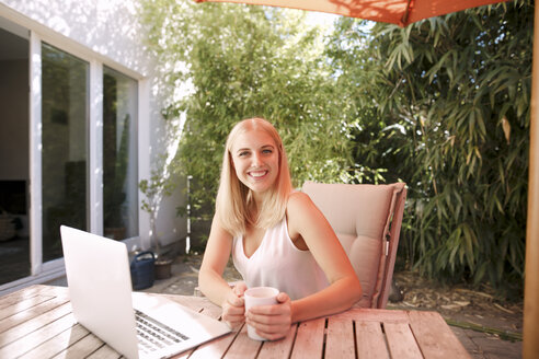 Junge lächelnde Frau, die einen Laptop benutzt und auf einem Gartentisch sitzt - KMKF00406