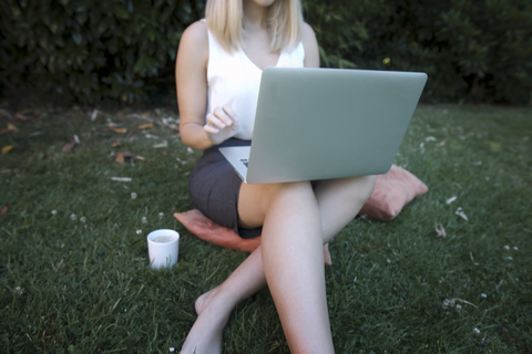 Junge Frau sitzt auf einem Kissen auf einer Wiese und benutzt einen Laptop, lizenzfreies Stockfoto