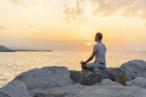 Spanien: Mann meditiert bei Sonnenaufgang am felsigen Strand - AFVF01064