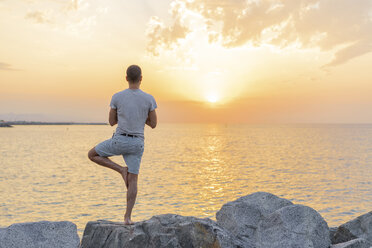 Spanien: Mann macht Yoga bei Sonnenaufgang am felsigen Strand, Baumposition, Rückansicht - AFVF01066