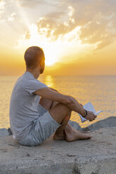 Spanien, Mann liest ein Buch bei Sonnenaufgang auf Felsen am Strand - AFVF01067