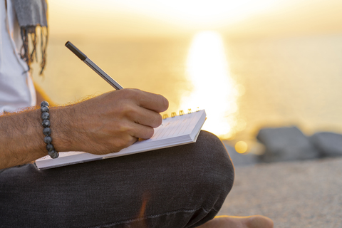 Spanien: Mann schreibt bei Sonnenaufgang auf einem Felsen am Strand in ein Notizbuch, lizenzfreies Stockfoto