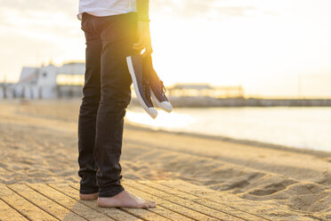 Spain. Man holding shoes, at a beach during sunrise - AFVF01071