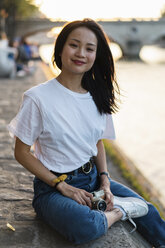 France, Paris, portrait of smiling young woman with camera at river Seine at sunset - AFVF01145