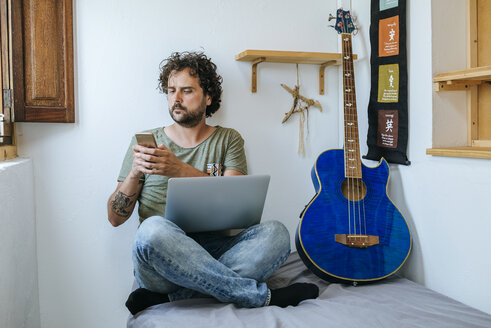 Mann in seinem Zimmer mit Mobiltelefon, Laptop und Bassgitarre - KIJF01973