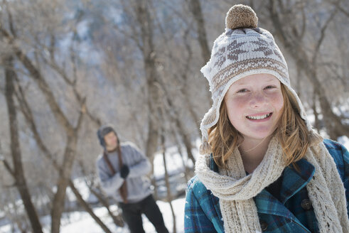 Winterlandschaft mit Schnee auf dem Boden. Ein junges Mädchen mit Bommelmütze und Schal im Freien. Ein Mann im Hintergrund. - MINF02953