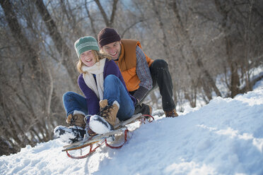 Winterlandschaft mit Schnee auf dem Boden. Ein Mann schiebt eine junge Frau auf einem Schlitten von einem Abhang hinauf. - MINF02955
