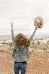 Rear view of a woman standing in the desert, holding a hat, her arms raised up in the air. - MINF02991